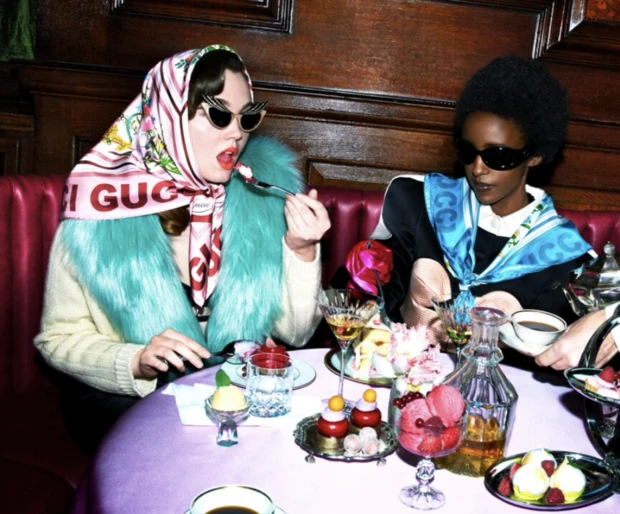 Two fashionable women wearing headscarves and sunglasses sit at a table covered with desserts and drinks in a restaurant setting.