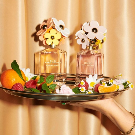 Two Marc Jacobs Daisy perfume bottles on a silver tray with fruit and flowers, held by a manicured hand with a gold bracelet.