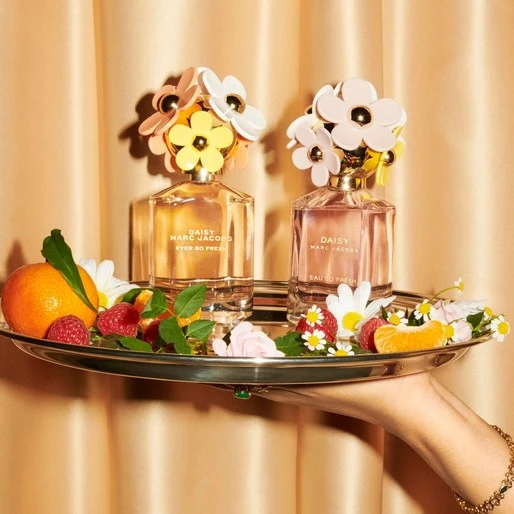 Two Marc Jacobs Daisy perfume bottles on a silver tray with fruit and flowers, held by a manicured hand with a gold bracelet.