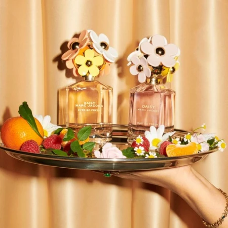 Two Marc Jacobs Daisy perfume bottles on a silver tray with fruit and flowers, held by a manicured hand with a gold bracelet.