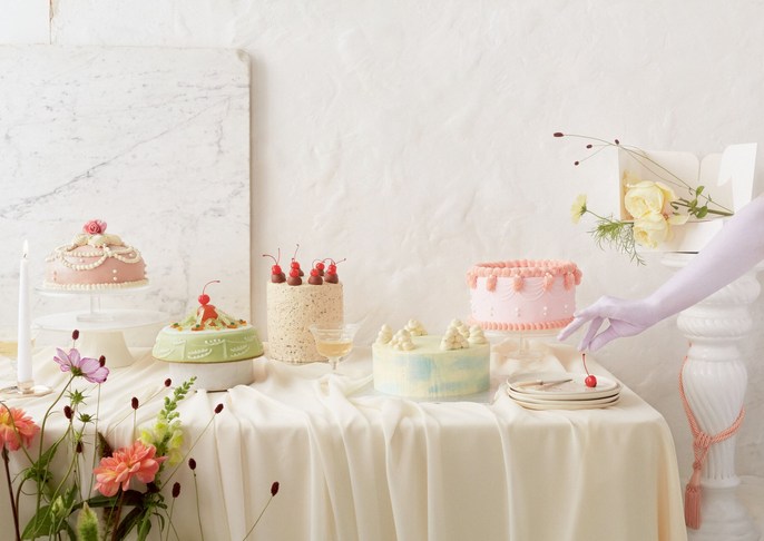 Variety of decorated cakes on a draped table, complemented by flowers and a hand placing a cherry on a plate. Soft, diffused lighting.