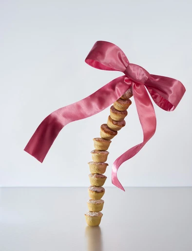Tower of miniature tarts stacked on a white reflective surface, accented with a decorative pink satin bow and long flowing ribbons.