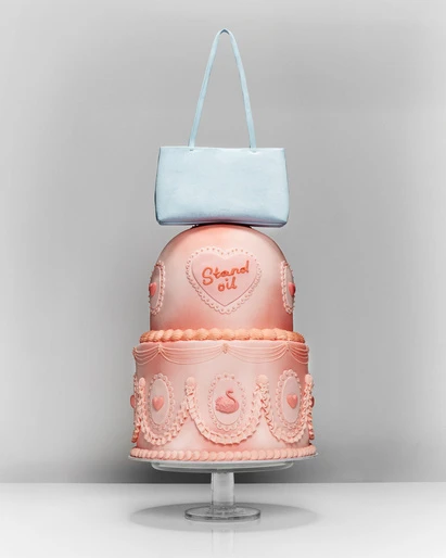 A three-tiered pink cake decorated with swans and hearts sits on a clear stand with a light blue handbag balanced on top of it.