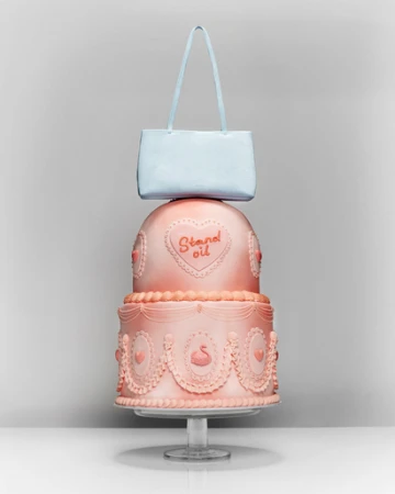 A three-tiered pink cake decorated with swans and hearts sits on a clear stand with a light blue handbag balanced on top of it.