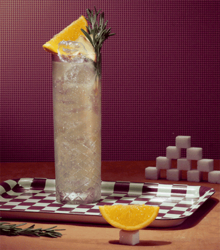 Tall glass of cocktail garnished with an orange slice and rosemary, set on a checkered tray with sugar cubes against a dark red background.