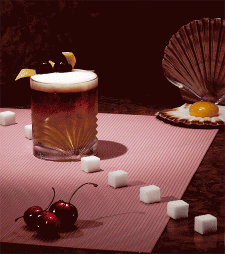 A vintage-style cocktail garnished with cherries and lemon peel sits on a pink surface with sugar cubes and a seashell.