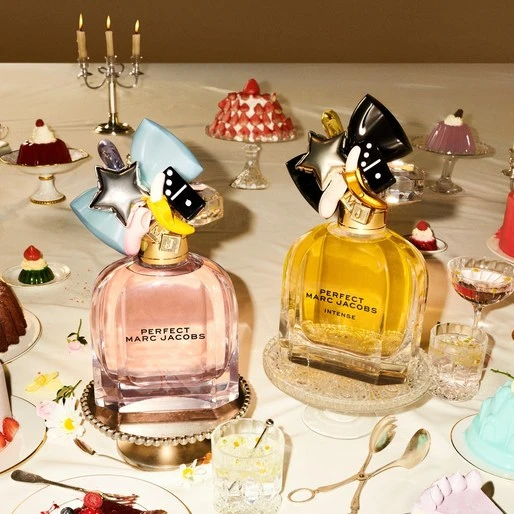 Two bottles of Marc Jacobs Perfect Intense perfume are displayed among cakes and desserts on a table with a light background.