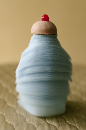 Abstract dessert-like sculpture in pale blue fabric with a pink cap and red cherry detail, set against a beige backdrop and textured surface.