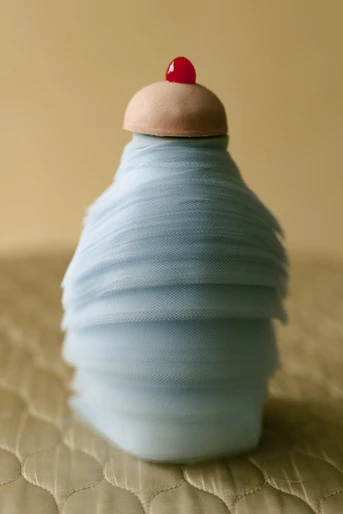 Abstract dessert-like sculpture in pale blue fabric with a pink cap and red cherry detail, set against a beige backdrop and textured surface.
