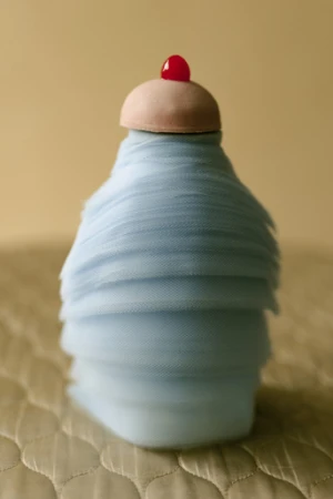 Abstract dessert-like sculpture in pale blue fabric with a pink cap and red cherry detail, set against a beige backdrop and textured surface.