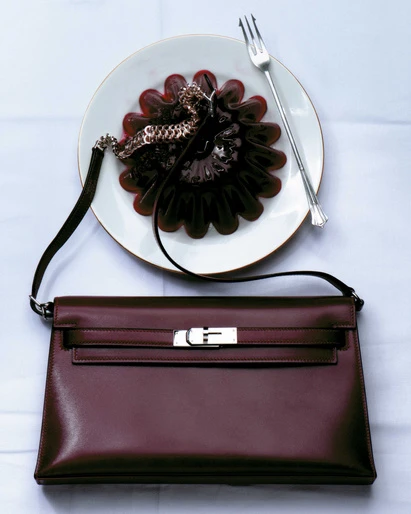 Burgundy leather purse on a white tablecloth, next to a white plate with red gelatin dessert and a silver fork in an indoor setting.