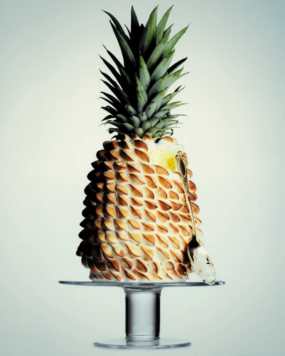 Close-up studio shot of a pineapple cake with meringue peaks on a clear cake stand with a metal server and a neutral background.