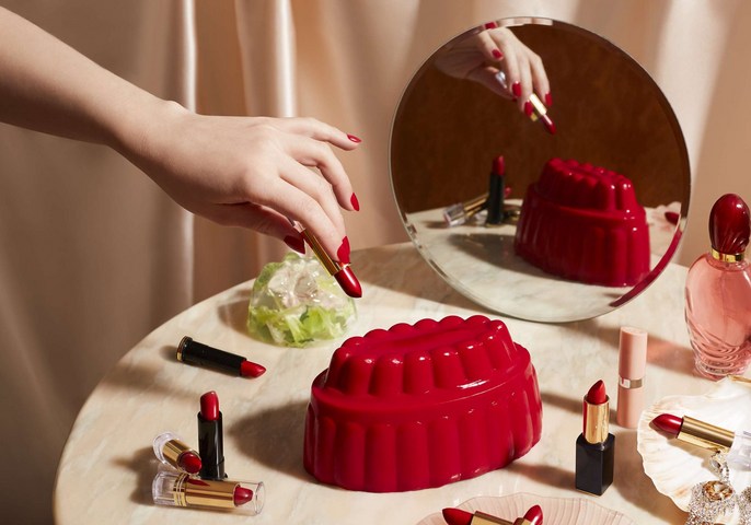 Close-up studio shot of a woman's hand with red nail polish reaching for a red lipstick next to several cosmetic items on a marble surface.