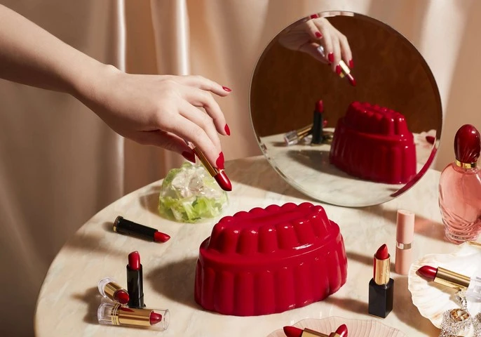 Close-up studio shot of a woman's hand with red nail polish reaching for a red lipstick next to several cosmetic items on a marble surface.