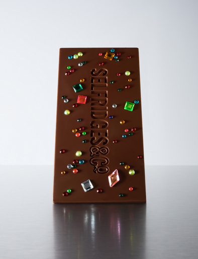Studio shot of a chocolate bar decorated with colorful gems and the embossed brand name 'Selfridges & Co.' against a gradient background.