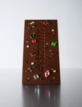 Studio shot of a chocolate bar decorated with colorful gems and the embossed brand name 'Selfridges & Co.' against a gradient background.
