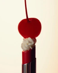 A vibrant red cherry with stem atop a dollop of whipped cream resting on a red lipstick tube
