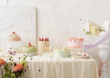 Multiple decorated cakes displayed on a draped table with flowers and a hand in lavender gloves placing a cherry