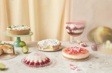 A colorful arrangement of assorted desserts, cakes, pies, and jellies styled on a light stone countertop with soft fabric drapes in the background.