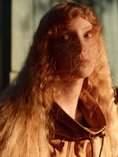 Portrait of a person with long, wavy, honey-blonde hair partially obscuring the face, wearing a tan-colored jacket, in soft outdoor lighting.
