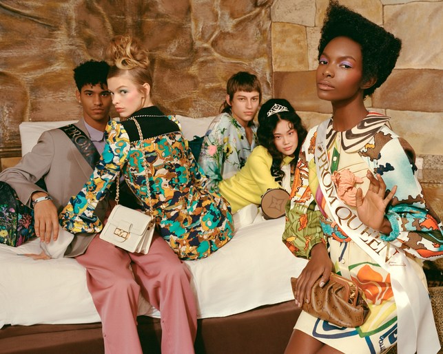 Group of five young adults in retro-style clothing pose indoors, one wears a 'Prom Queen' sash, with vintage Louis Vuitton bags.