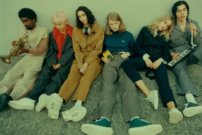 Group of six young adult musicians sitting in casual apparel. Holding trumpet, keyboard, and music player in a neutral indoor setting.