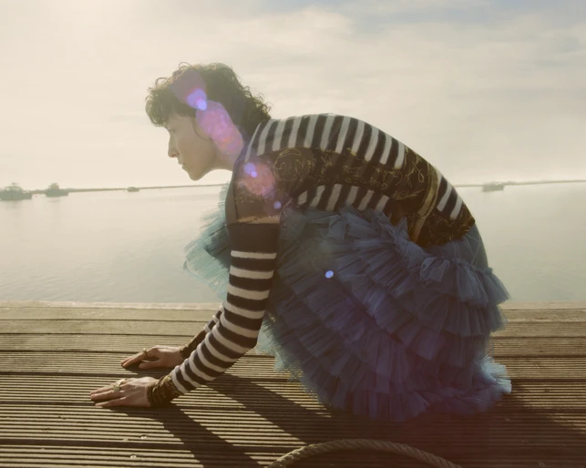 Adult female in a black and white striped top and blue tulle skirt crouches on a wooden dock near a body of water, with a cloudy sky above.
