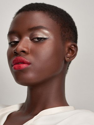 Close-up beauty shot of a dark-skinned woman with short hair, wearing bright red lipstick and gold eyeshadow against a plain background.