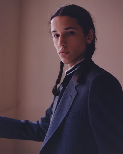 Portrait of a young adult with braided dark hair wearing a dark blue suit jacket and black and white striped shirt against a warm-toned background.