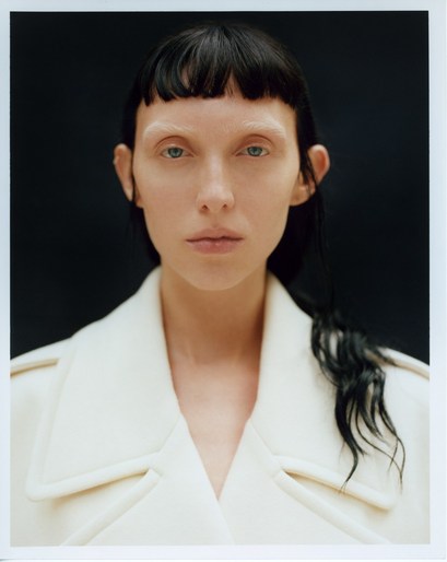Close-up portrait of a woman with a white coat, dark hair, and light skin in front of a dark background. Minimalist beauty shot.