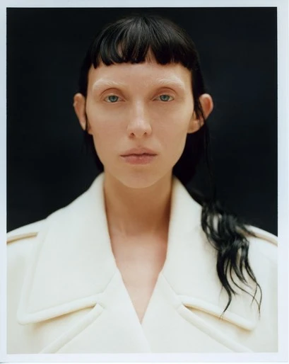 Close-up portrait of a woman with a white coat, dark hair, and light skin in front of a dark background. Minimalist beauty shot.