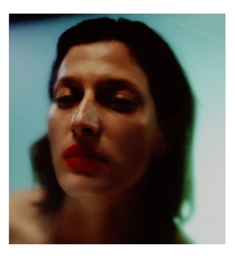 Close-up portrait of an adult woman with dark hair and red lipstick against a blurred light blue background in a studio setting.