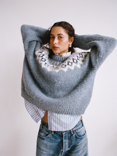Adult woman poses in a grey sweater and jeans against a white background, with her hands behind her head and natural expression.