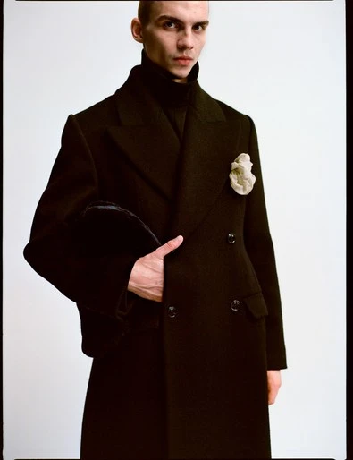 Portrait of a slim adult male fashion model in a dark, double-breasted overcoat holding a black hat against a plain white background.
