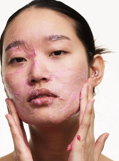 Close-up portrait of an Asian woman with pink cream applied to her face, hands gently touching her cheeks, beauty treatment.
