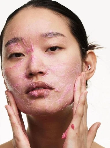 Close-up portrait of an Asian woman with pink cream applied to her face, hands gently touching her cheeks, beauty treatment.