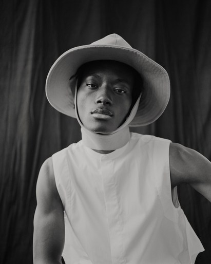 Portrait of a young man in a white sleeveless shirt and wide-brimmed hat against a dark backdrop in a photo studio with natural lighting.