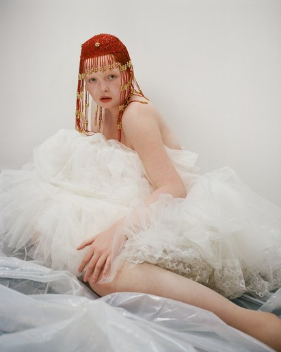 A fair-skinned person with red hair and a beaded headpiece reclines in a puffy white gown against a white wall and grey surface.