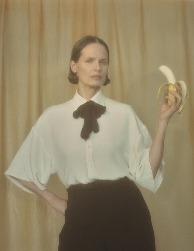 A woman with a short bob haircut poses indoors, holding a peeled banana against a light yellow backdrop, wearing a white blouse and dark skirt.