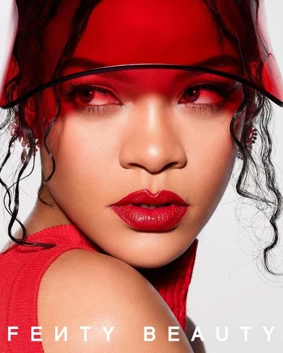 Close-up of a woman with red lipstick and a translucent red visor, posing for Fenty Beauty against a neutral background.