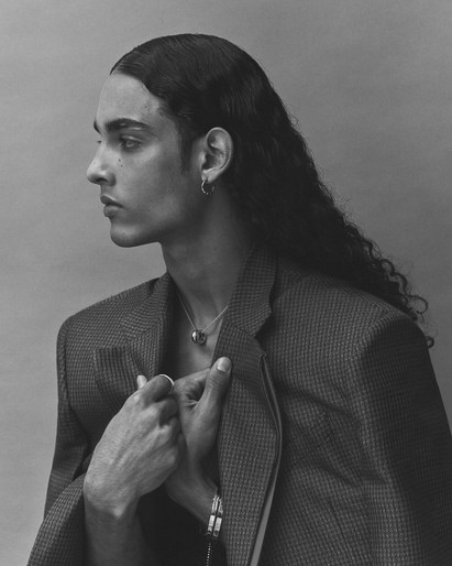 Profile portrait of a stylish adult male with long, dark, wavy hair wearing a blazer jacket and jewelry against a plain backdrop.