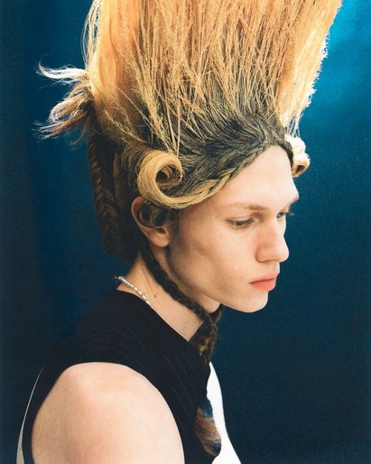Portrait of a young adult with an elaborate blonde hairstyle, wearing a black tank top, set against a dark blue backdrop.
