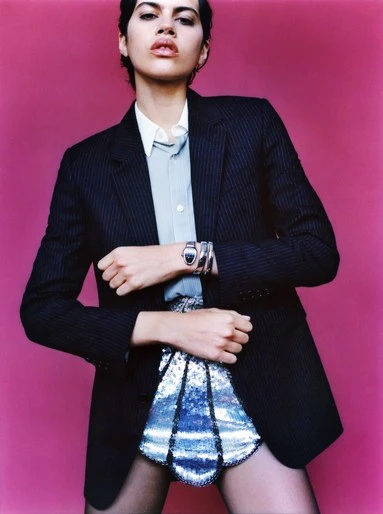 Woman poses in blazer and sequin mini-skirt against a pink background. She wears bracelets, a watch and a light-blue collared shirt.