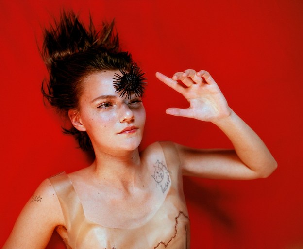 Portrait of a woman with tousled hair, dark eye decoration, and a neutral expression against a vibrant red background.