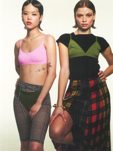 Two women pose in a photo studio setting, wearing coordinated plaid skirt and spandex styles, showcasing 90s-inspired fashion trends.