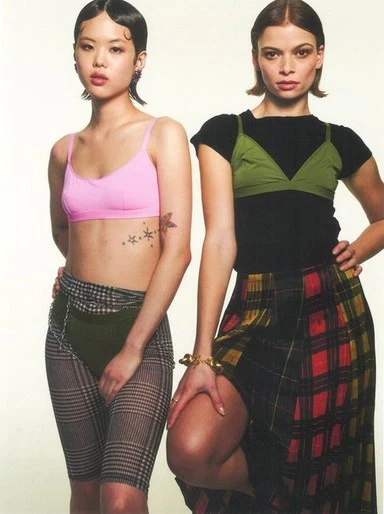 Two women pose in a photo studio setting, wearing coordinated plaid skirt and spandex styles, showcasing 90s-inspired fashion trends.