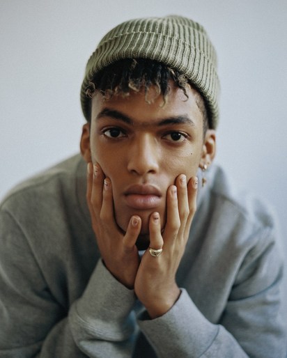 Close-up studio portrait of a young adult male with short curly hair wearing a gray sweatshirt and beanie with hands on face.