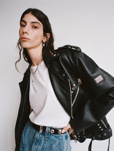 Woman in a black leather jacket, white T-shirt, and blue jeans stands against a white background in a fashion shot.