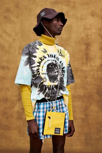 Black male model posing in yellow and blue plaid shorts, tie-dye t-shirt, yellow turtleneck, brown hat, and a yellow purse against brown backdrop.