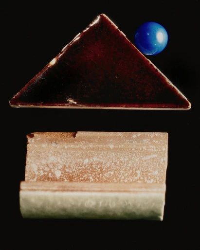Abstract still life featuring a dark brown triangle tile, a rectangular tile, and a blue sphere against a black backdrop.
